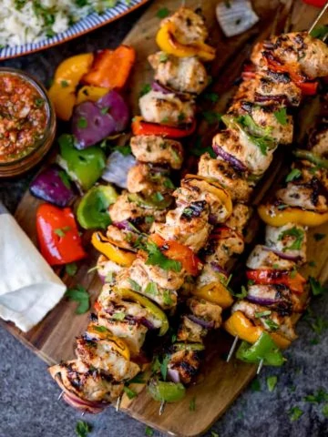 Square overhead shot of Mexican chicken kebabs piled on a wooden board. Salsa and rice in the background.