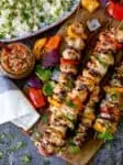 Mexican chicken kebabs piled on a wooden board. Salsa and rice in the background.