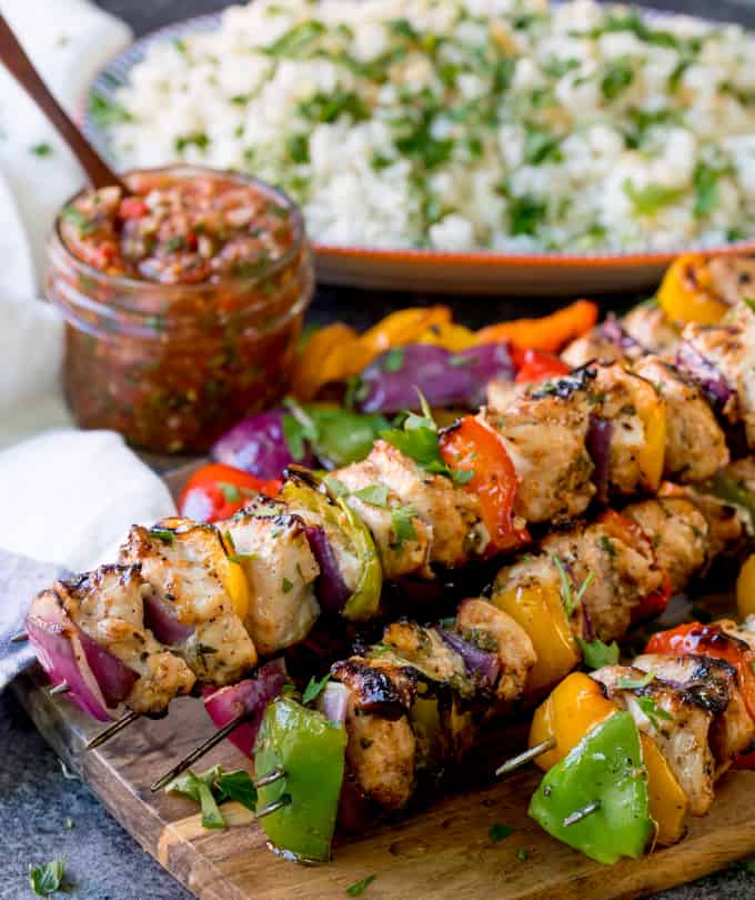 Close up image of Mexican chicken kebabs piled on a wooden board. Salsa and rice in the background.