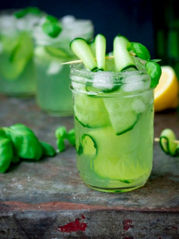 Square image of gin, cucumber and basil cocktails in mason jars, on a metal tray. There are ingredients for the cocktail scattered on the tray.