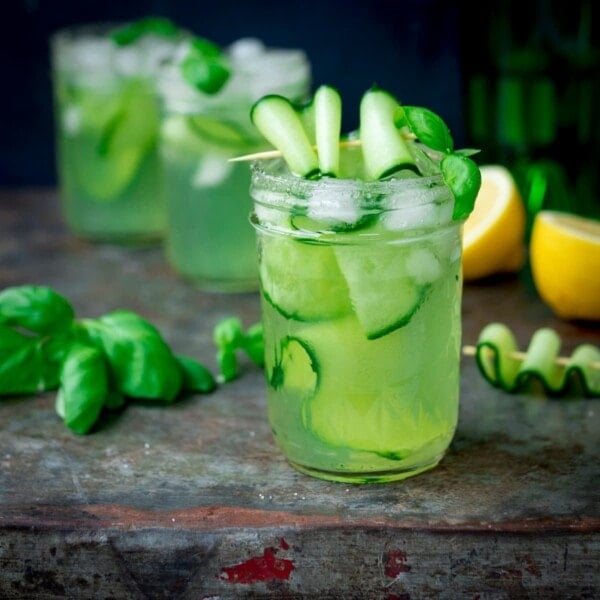 Square image of gin, cucumber and basil cocktails in mason jars, on a metal tray. There are ingredients for the cocktail scattered on the tray.