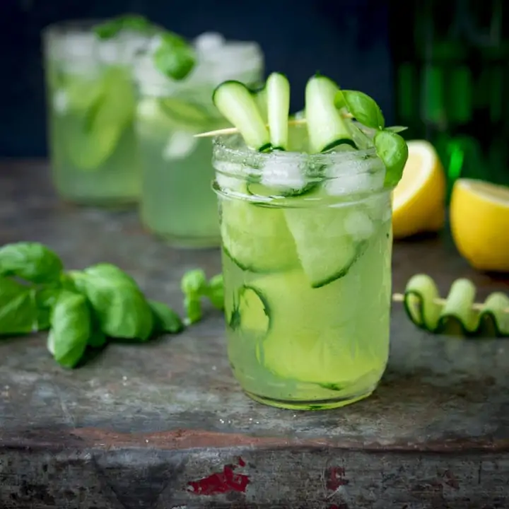 Square image of gin, cucumber and basil cocktails in mason jars, on a metal tray. There are ingredients for the cocktail scattered on the tray.