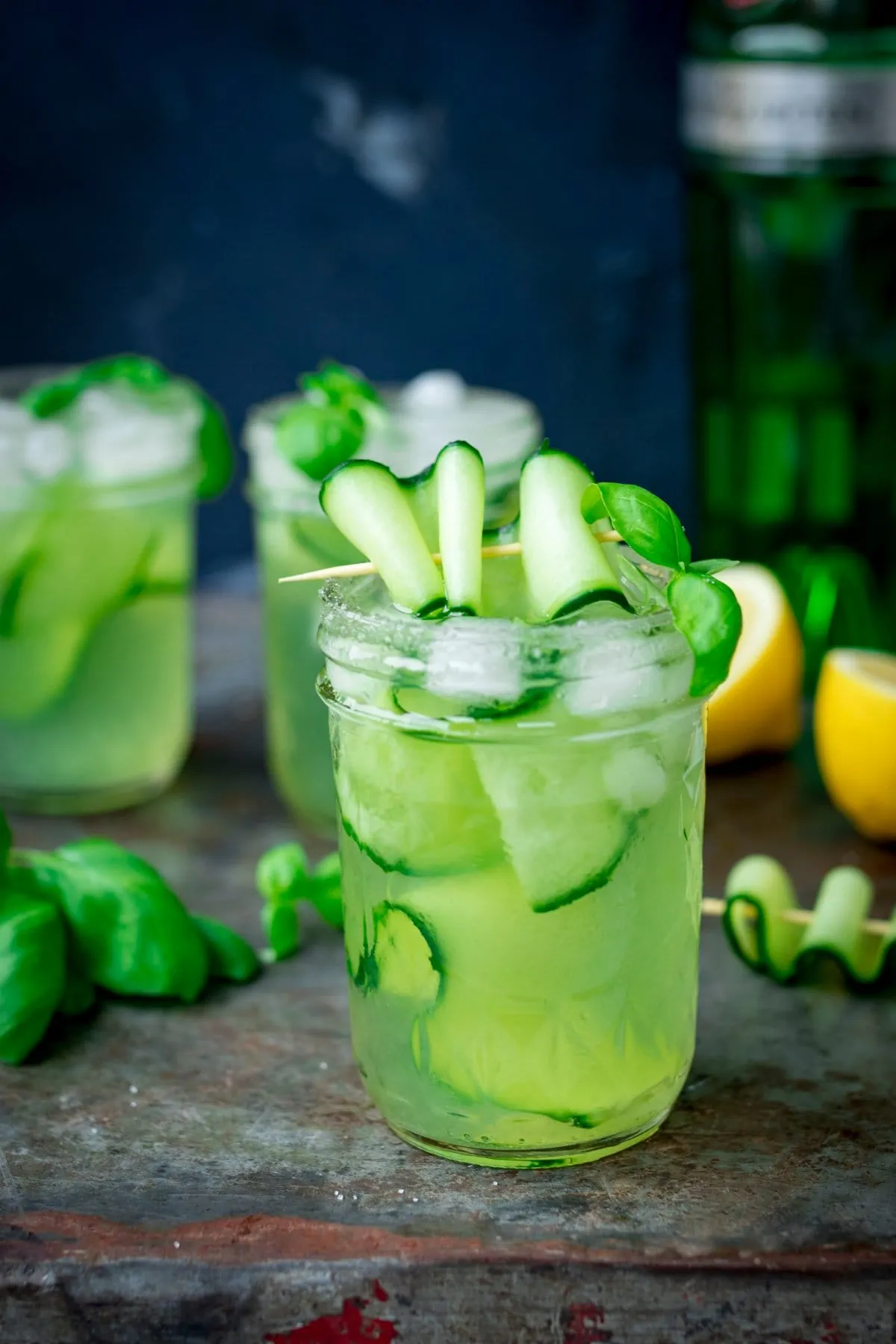 Tall image of gin, cucumber and basil cocktails in mason jars, on a metal tray. There are ingredients for the cocktail scattered on the tray.