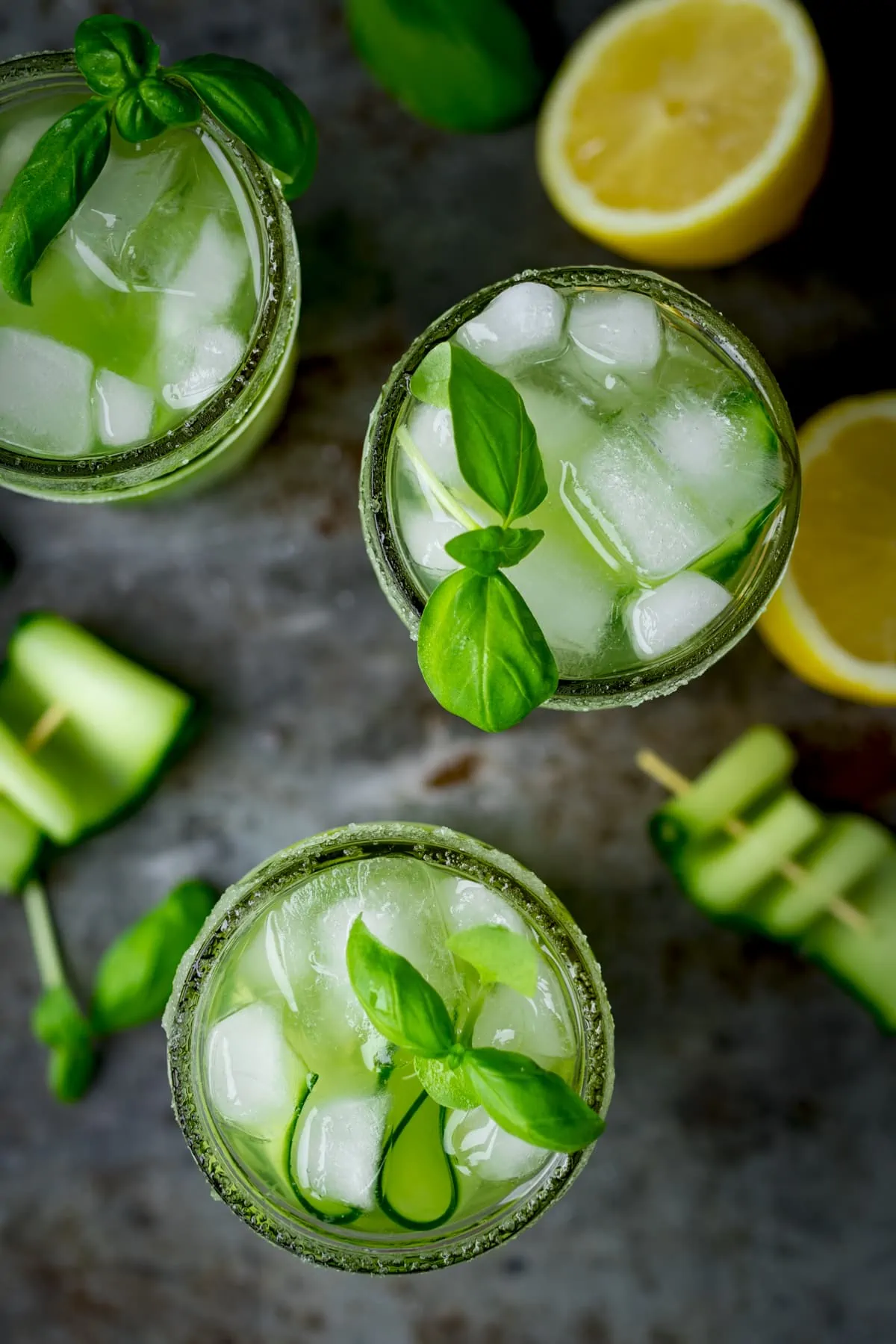 Overhead image of three mason jars filled with gin, cucumber and basil cocktails. There are cucumber ribbons and sliced lemons on the tray around the cocktails.