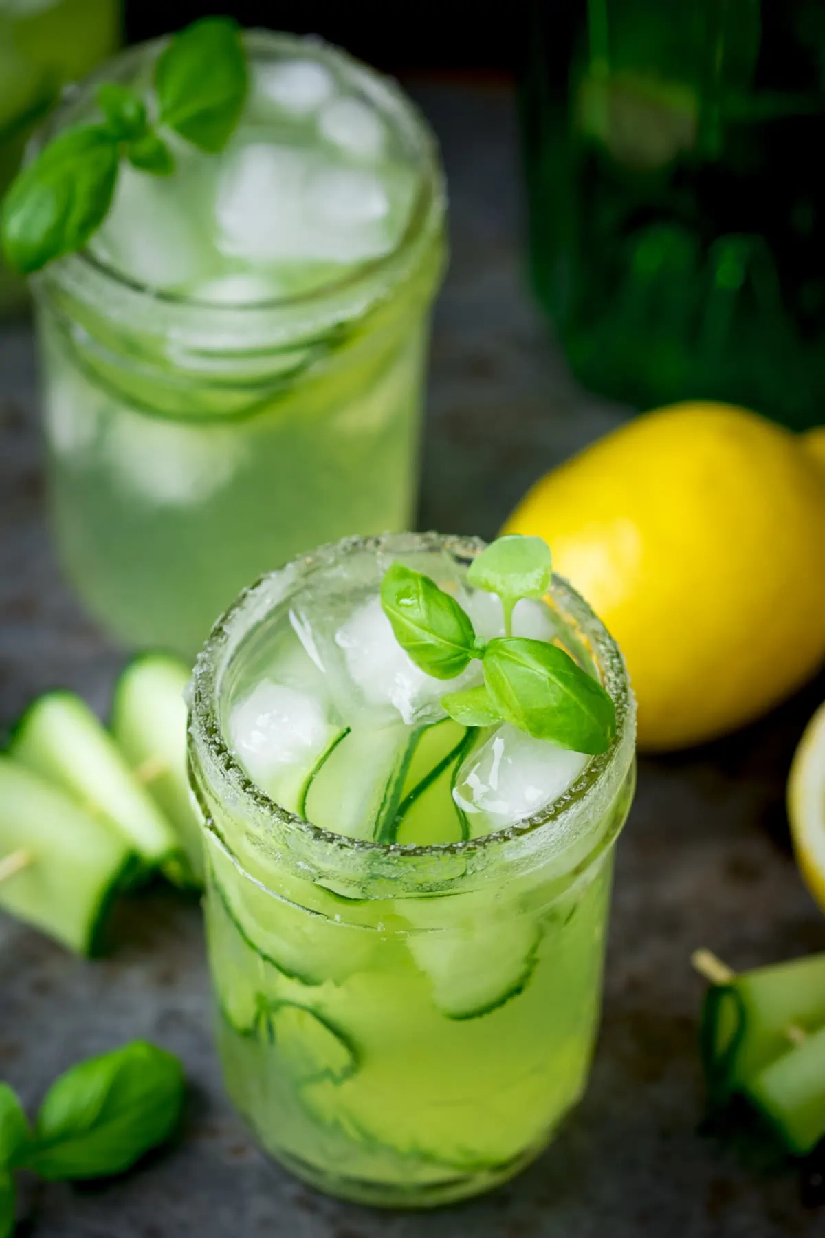 Two gin, cucumber and basil cocktails in mason jars with a sugar rim. There are ingredients scattered around the glasses.