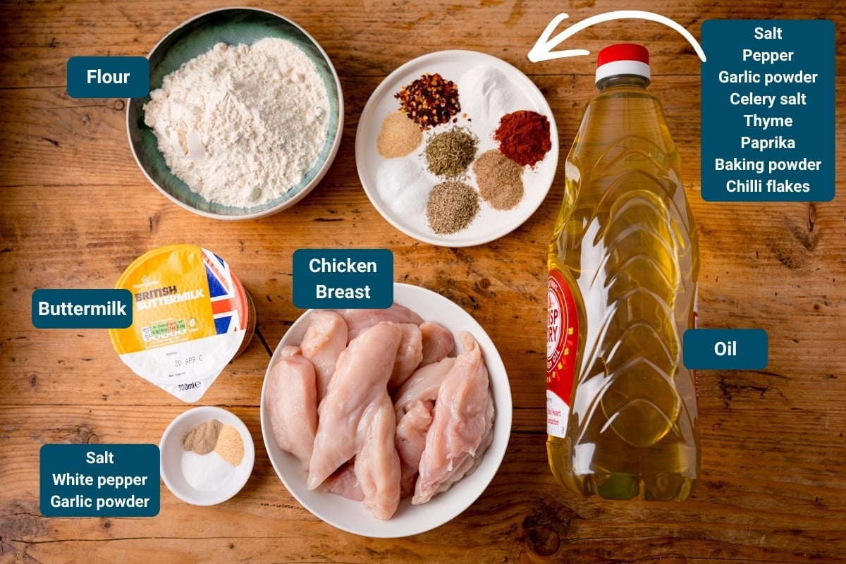 Ingredients for Crispy Chicken tenders are laid out on a wooden surface, including: flour, buttermilk, chicken breast, oil, and various labelled spices in bowls. All ingredients are labelled.