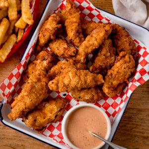 Overhead image of a tray of crispy chicken tenders with chicken tender dipping sauce on red checkered paper, on a tray, served with golden crinkle-cut chips.