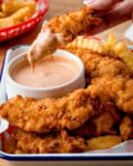 A person dips a Crispy Chicken Tender into chicken-tender-dipping sauce, with golden crinkle-cut chips in the background.
