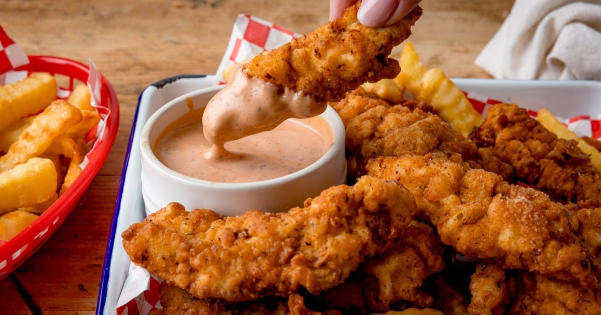 A hand dips a Crispy Chicken tender into a bowl of dipping sauce, while more chicken tenders and crinkle-cut chips fries are on trays in the background.