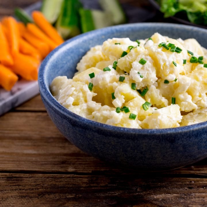 Creamy potato salad in a blue bowl on a wooden table. Chopped vegetables in the background.