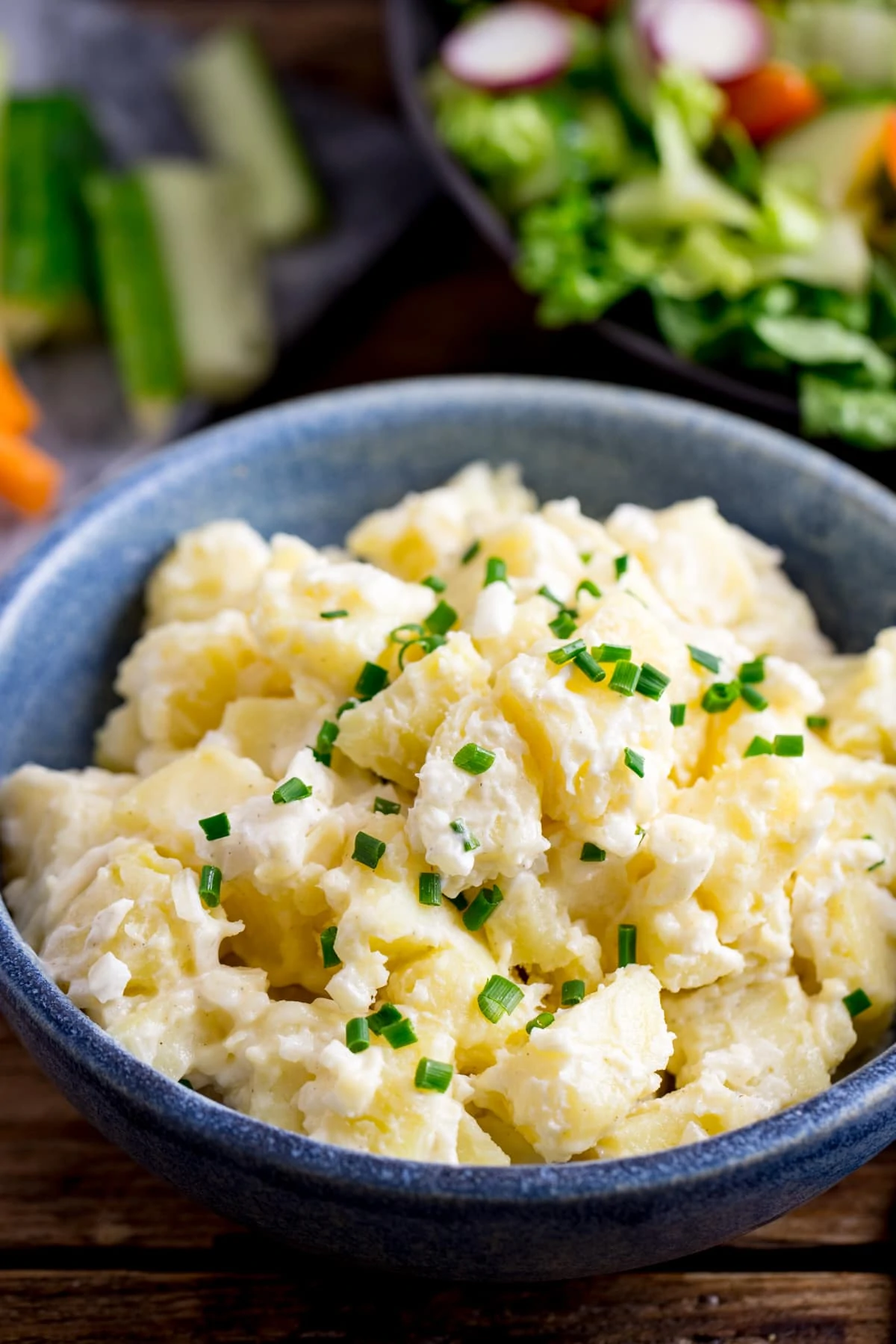 Creamy potato salad in a blue bowl. Chopped vegetables and a bowl of salad in the background.