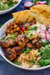 spicy chicken, chickpeas, couscous, leaves, radish, hummus and flatbreads in a bowl