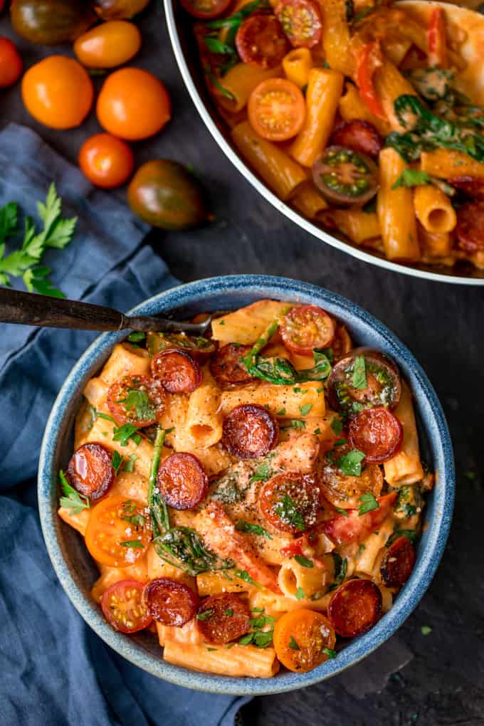 Overhead shot of a blue bowl filled with tomato and chorizo rigatoni on a blue surface next to a blue napkin. There is a pan of the pasta dish in the top of the frame. There are cherry tomatoes and fresh parsley next to the bowl.