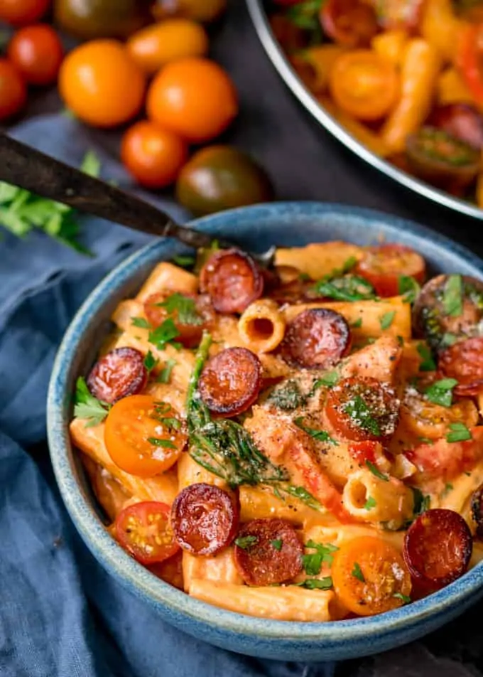 Creamy Tomato and Chorizo Rigatoni with spinach and tomatoes in a blue bowl on a blue surface, next to a blue napkin. There are tomatoes and parsley also in shot.