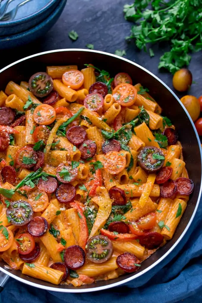 One Pot Creamy Tomato and Chorizo Rigatoni in a pan with spinach and tomatoes on a blue surface. There are tomatoes and fresh parsley scattered around the pan