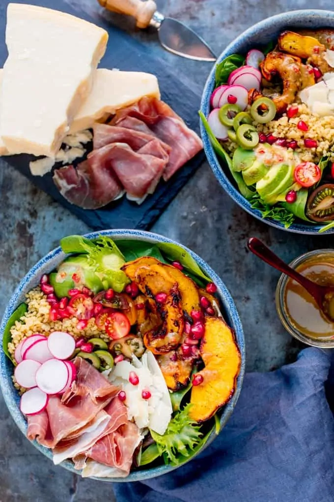 Overhead Photo of 2 bowls of Winter Nourish Bowl filled with goodies like roasted pumpkin, bulgur wheat and avocado with some Grana Padano and Prosciutto on a slate board
