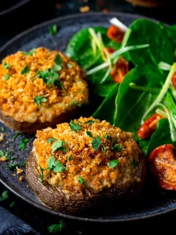 Square image of Stuffed mushrooms on a black plate with salad