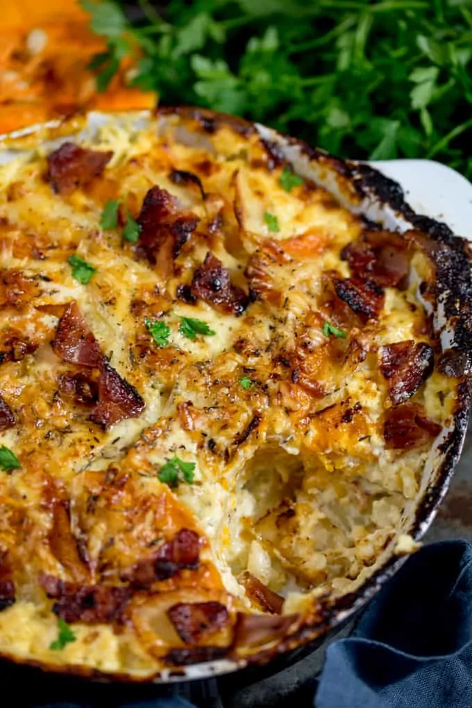 Close up Photo of Squash and Potato Gratin in a grey and white backing dish with a scoop taken out, topped with bacon and parsley in the background