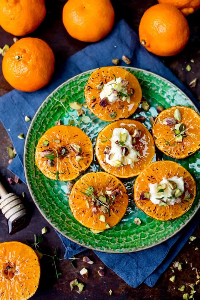 Overhead picture of Brown Sugar Glazed Clementines with Cream Cheese, Honey and Pistachios on a green plate sat on a blue cloth surrounded by whole clementines
