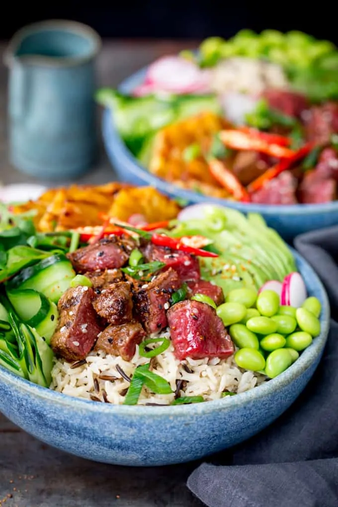 Side on shot of steak poke bowl with rice, cucumber and edamame in a blue bowl. Further poke bowl in the background.