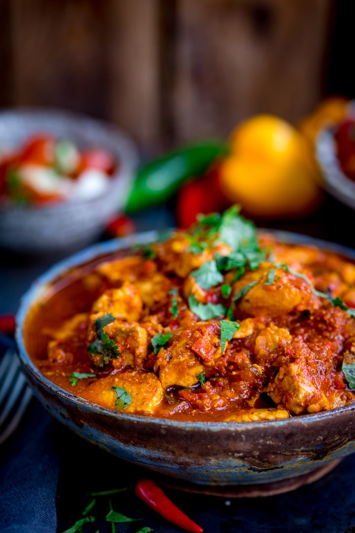 Close up photo of Healthier Slow Cooked Spicy Chicken Rogan Josh in a stone bowl on a blue board with chilli peppers in the background