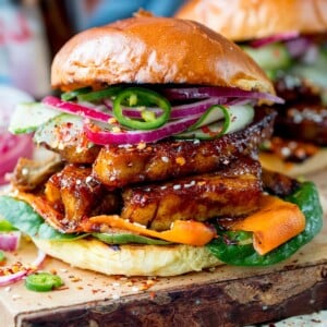 Square side-on image of toasted brioche roll filled with sticky pork belly strips and pickled carrot, red onion and cucumber strips. Topped with jalapeno slices. Served on a wooden board.