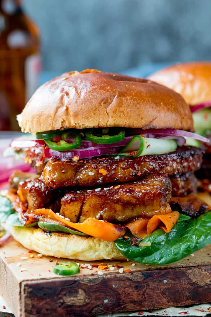 Side on shot of toasted brioche roll filled with sticky pork belly strips and pickled carrot, red onion and cucumber strips. Topped with jalapeno slices. Served on a wooden board.