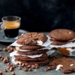 Close up a stack of double chocolate nougat cookies, with baking paper in between the cookies. Chocolate chips and a cookie with a bite taken out in foreground. Glass cup of espresso and further stack of cookies in background.