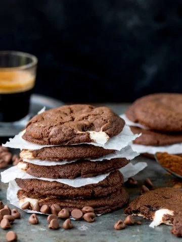 Close up a stack of double chocolate nougat cookies, with baking paper in between the cookies. Chocolate chips and a cookie with a bite taken out in foreground. Glass cup of espresso and further stack of cookies in background.