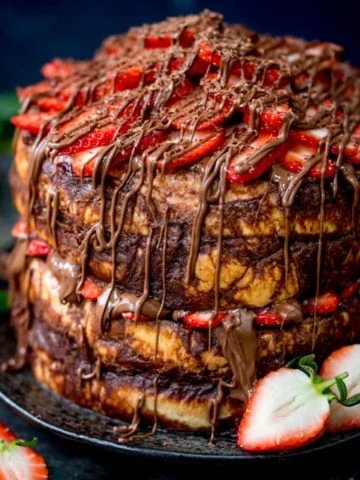 Close up image of a four layer chocolate bread and butter pudding round cake on a black plate. Nutella and strawberries on the top and in the middle. Dark blue background, strawberries scattered around.