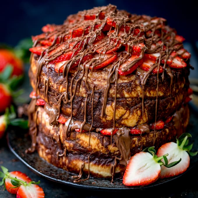Close up image of a four layer chocolate bread and butter pudding round cake on a black plate. Nutella and strawberries on the top and in the middle. Dark blue background, strawberries scattered around.