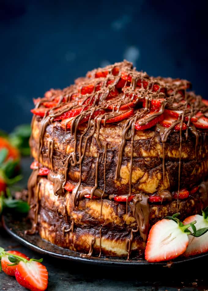 Tall image of a four layer chocolate bread and butter pudding round cake on a black plate. Nutella and strawberries on the top and in the middle. Cake is slightly cropped on the right hand side. Dark blue background, strawberries scattered around.