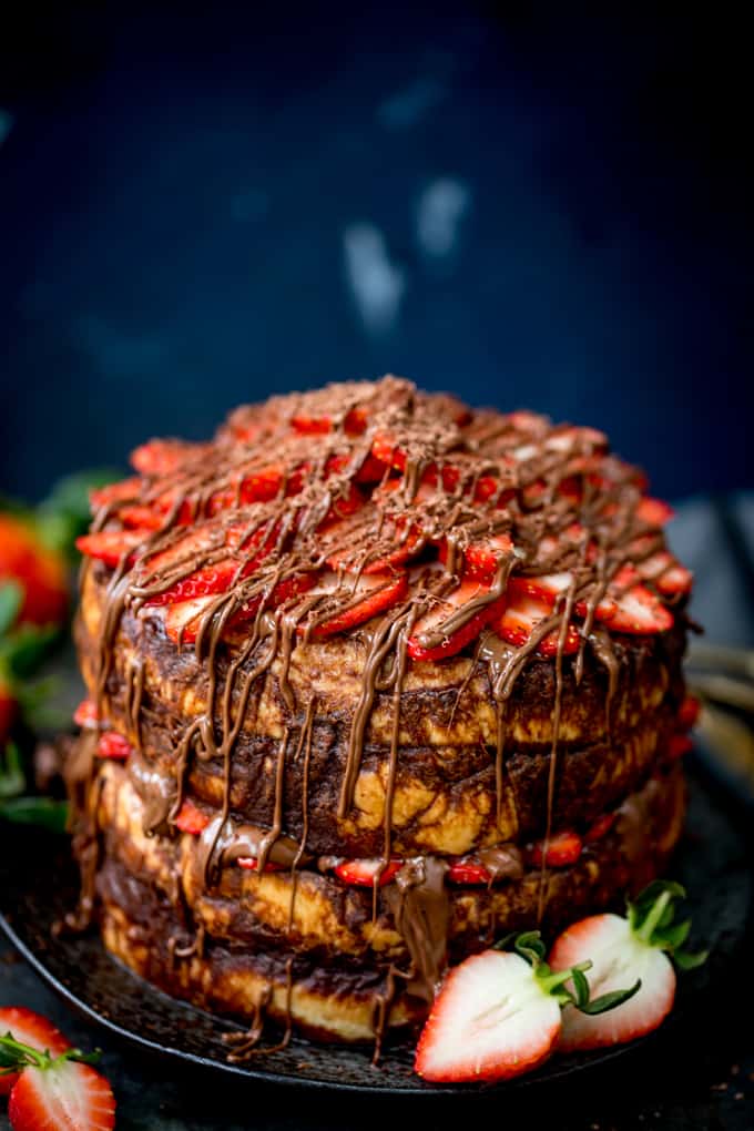 Tall image of a centre-aligned four layer chocolate bread and butter pudding round cake on a black plate. Nutella and strawberries on the top and in the middle. Dark blue background, strawberries scattered around.