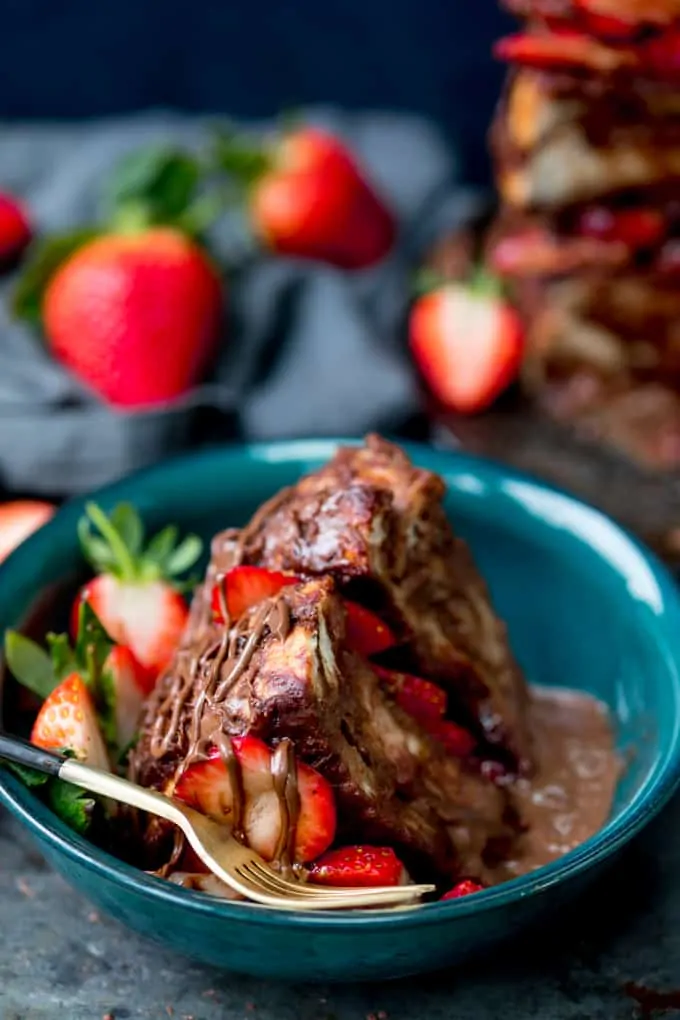 Slice of chocolate bread and butter pudding cake filled and topped with Nutella and strawberries in a dark green bowl with a gold fork with black handle. Drizzled with Nutella. Scattered strawberries and navy blue linen napkin in the background.