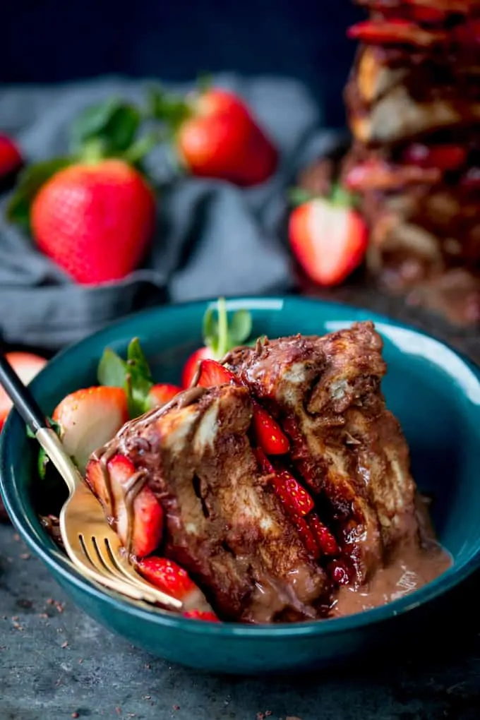 Slice of chocolate bread and butter pudding cake filled and topped with Nutella and strawberries in a dark green bowl with a gold fork with black handle. Scattered strawberries and navy blue linen napkin in the background.