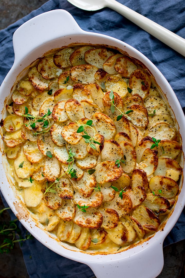 Overhead portrait photo of Creamy Chicken and Potato Bake with extra veggies in a grey and white casserole, topped with sprigs of thyme on a blue cloth