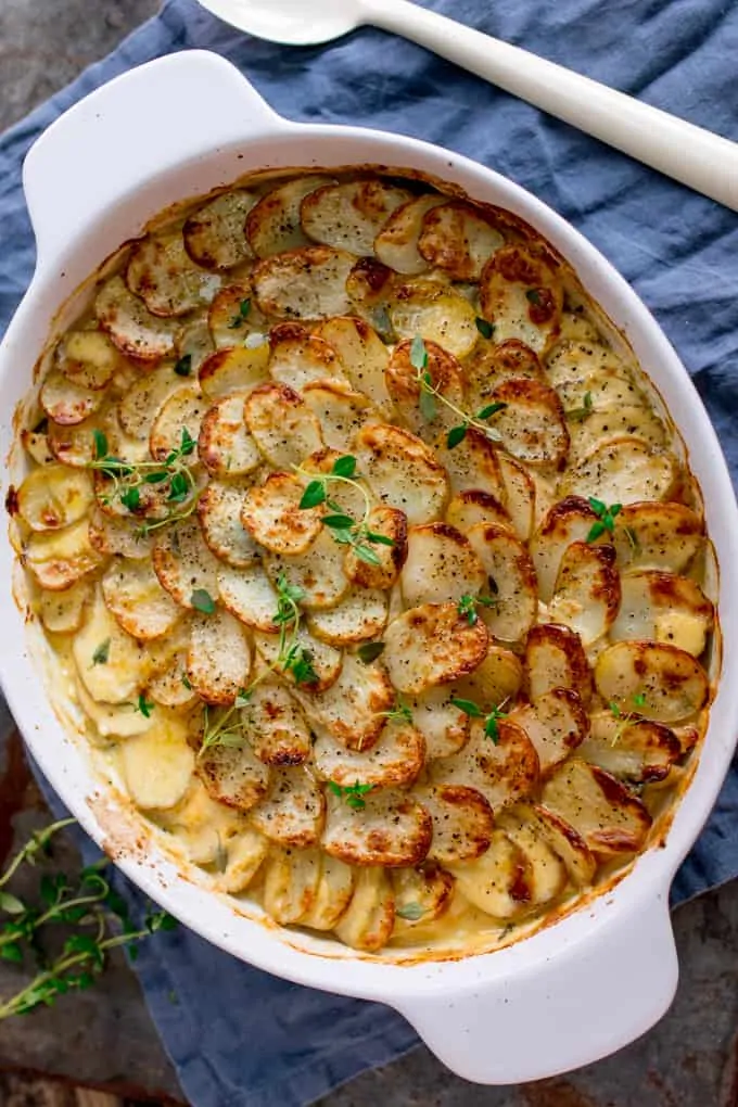 Overhead portrait photo of Creamy Chicken and Potato Bake with extra veggies in a grey and white casserole, topped with sprigs of thyme on a blue cloth