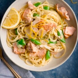 Square overhead image of salmon and spaghetti in a creamy lemon sauce in a white bowl with a lemon wedge in the bowl. The bowl is on a blue background. There is a fork and a napkin nearby.