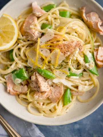 Square overhead image of salmon and spaghetti in a creamy lemon sauce in a white bowl with a lemon wedge in the bowl. The bowl is on a blue background. There is a fork and a napkin nearby.
