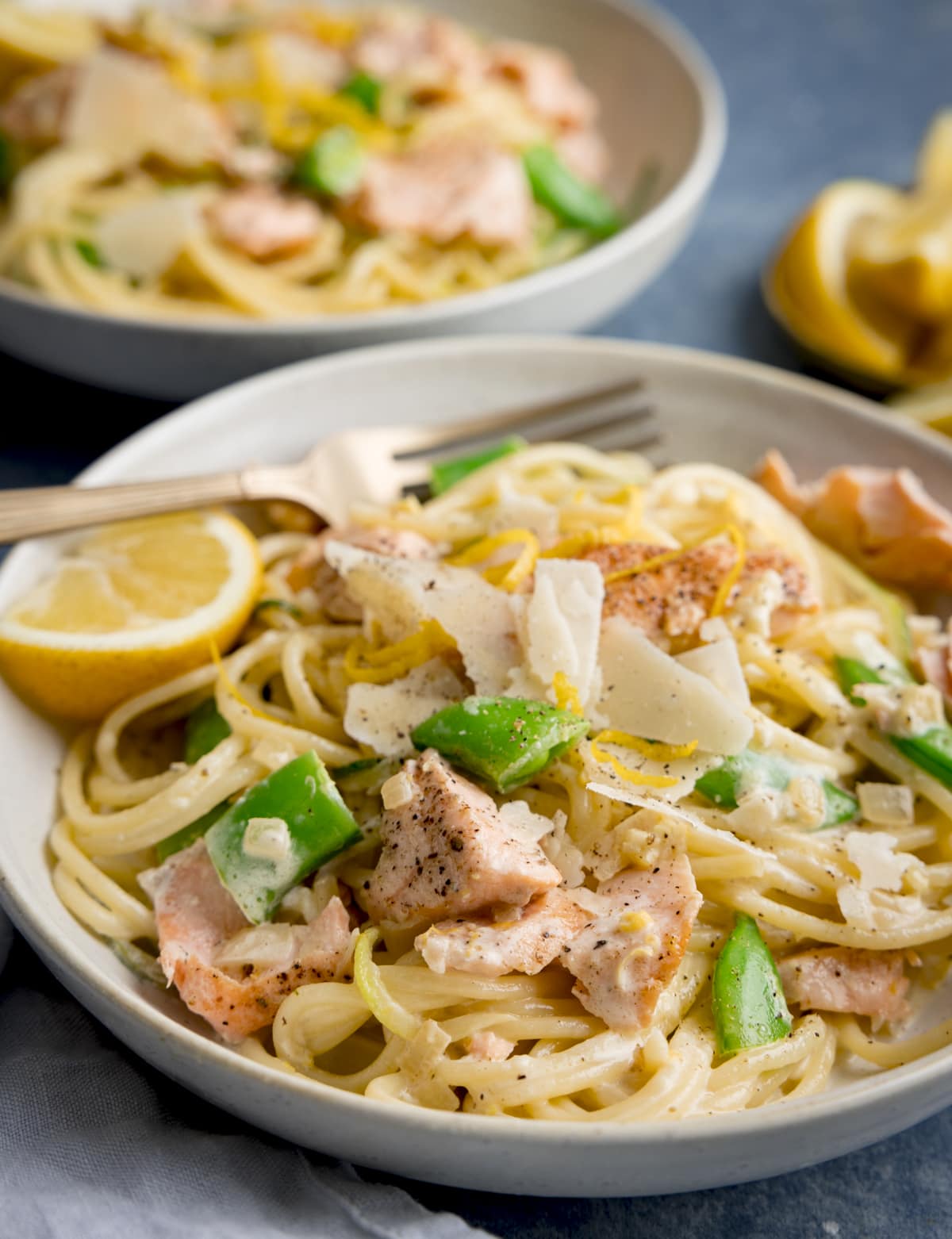 Side on image of salmon and spaghetti in a creamy lemon sauce in a white bowl with a lemon wedge in the bowl. The bowl is on a blue background. There is a further bowl of pasta, a napkin and some lemon wedges nearby.
