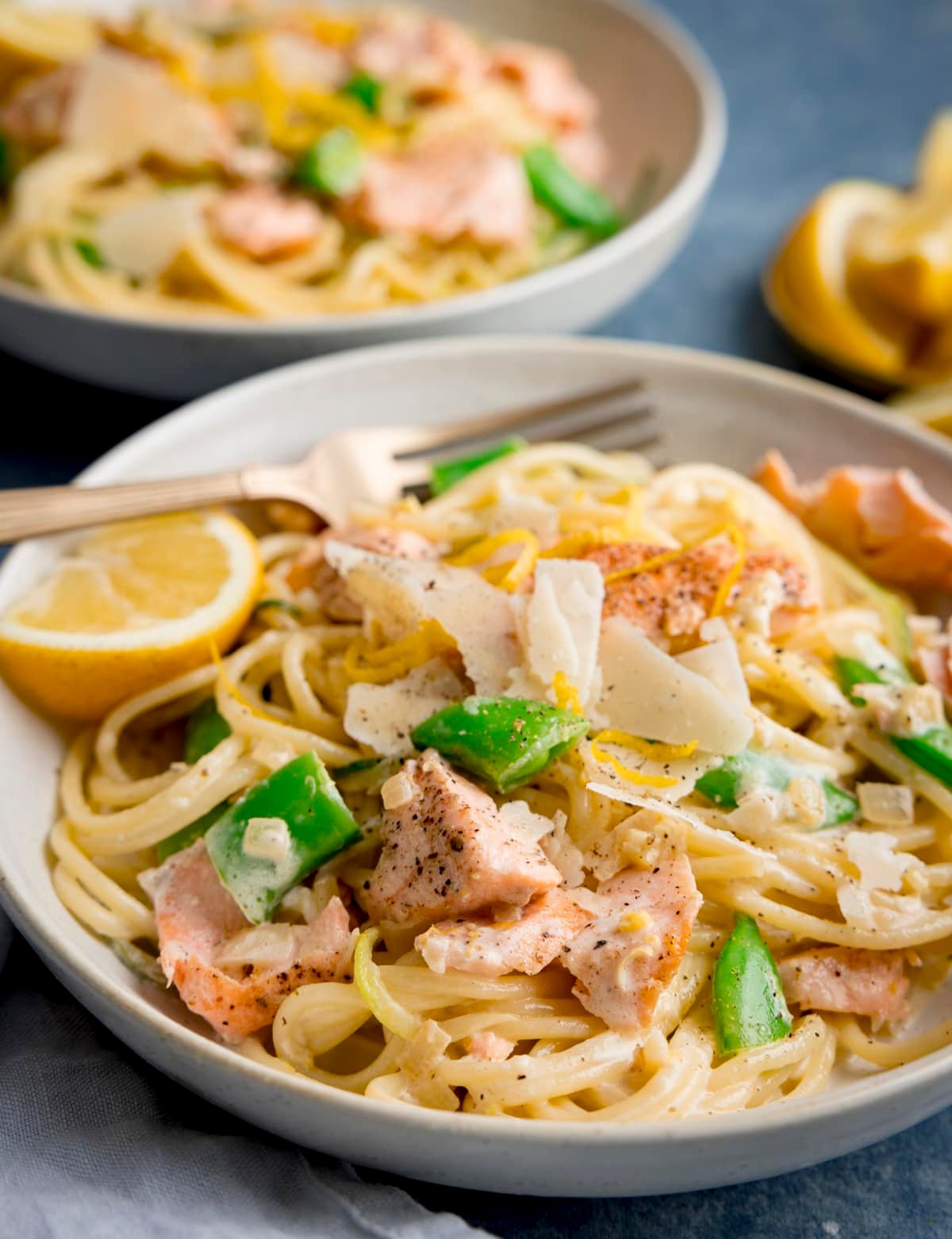 Side on image of salmon and spaghetti in a creamy lemon sauce in a white bowl with a lemon wedge in the bowl. The bowl is on a blue background. There is a further bowl of pasta, a napkin and some lemon wedges nearby.