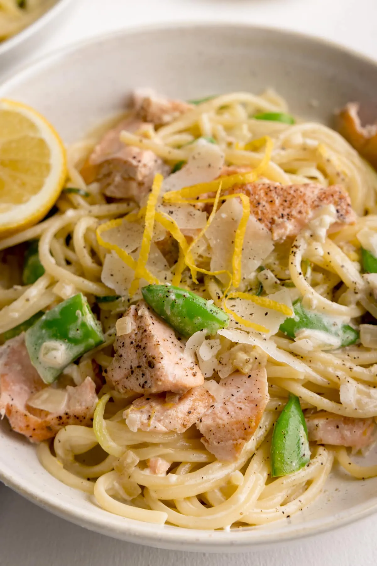 Close up of salmon and spaghetti in a creamy lemon sauce in a white bowl with a lemon wedge in the bowl. The bowl is on a white background.