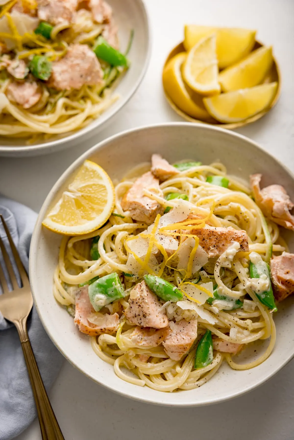 Overhead of salmon and spaghetti in a creamy lemon sauce in a white bowl with a lemon wedge in the bowl. The bowl is on a white background next to a small dish of lemon wedges. There is a further bowl of pasta, a fork and a napkin nearby.