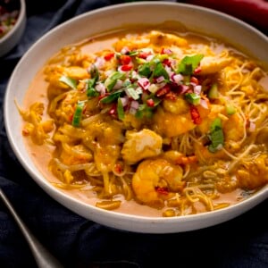 Square image of seafood laksa in a white bowl on a dark surface. The laksa is topped with a garnish of coriander, chili and red onion.