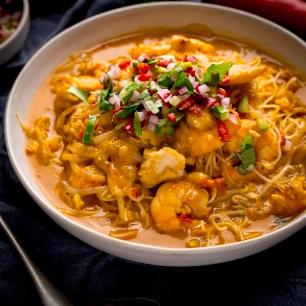 Square image of seafood laksa in a white bowl on a dark surface. The laksa is topped with a coriander-chilli-red onion garnish.