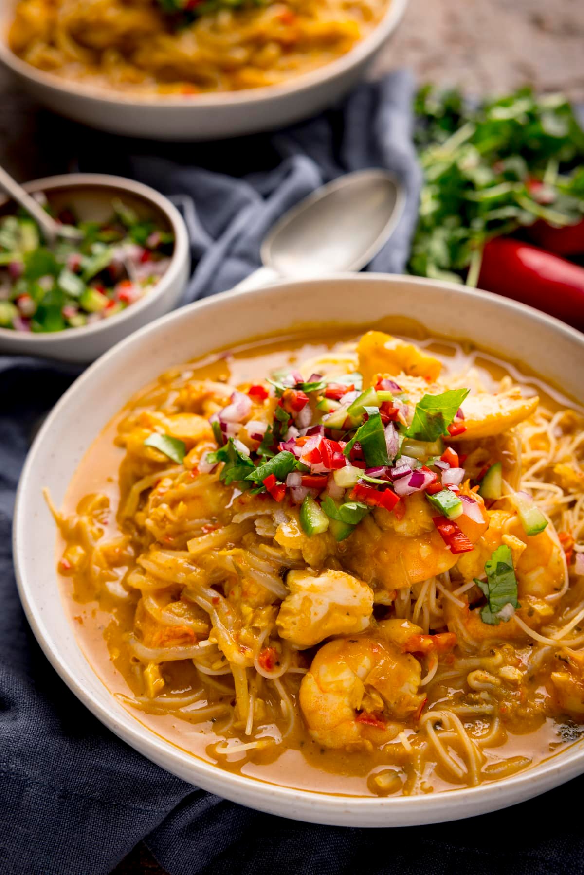 Seafood laksa in a white bowl on a wooden table. There is a napkin, a spoon and ingredients scattered around.
