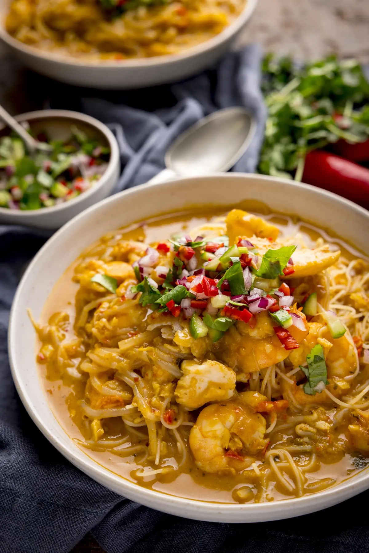 Seafood laksa in a white bowl, on a wooden table. There is a napkin, spoon and ingredients scattered around.