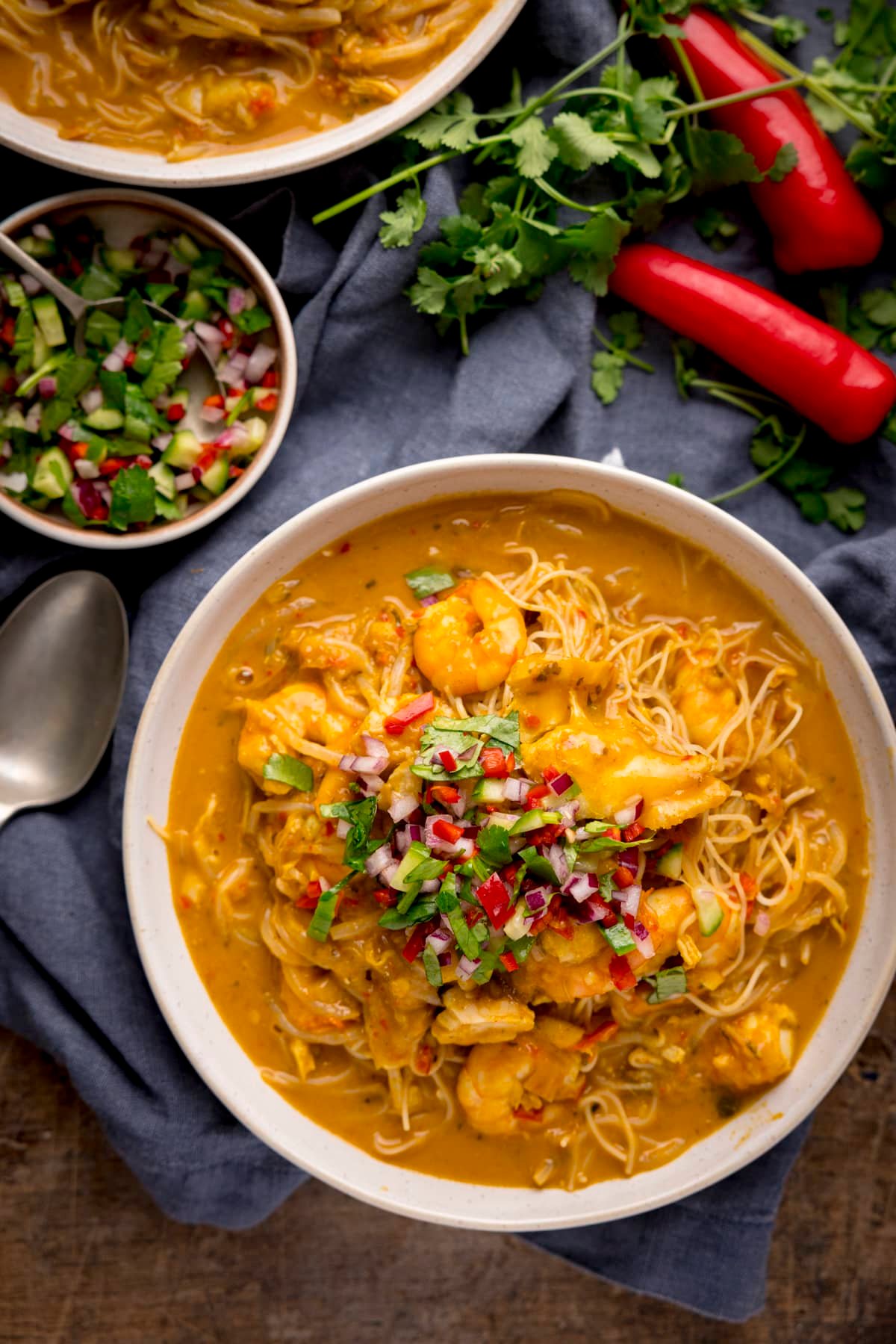 Overhead image of seafood laksa in a white bowl on a wooden table. There is a spoon, blue napkin, a garnish bowl, a second bowl of laksa and some chillies and coriander around the main bowl.