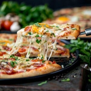 Side-on image of egg-topped breakfast pizza slice being lifted from the pizza. Melted cheese is stretching between the pizza and pizza slice.