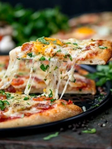 Side-on image of egg-topped breakfast pizza slice being lifted from the pizza. Melted cheese is stretching between the pizza and pizza slice.